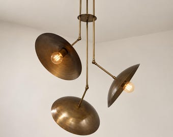 Può includere: Un lampadario in ottone con tre bracci regolabili che si estendono da un'asta verticale centrale. Ogni braccio sostiene un ampio paralume a forma di ciotola con una lampadina a luce calda al centro, creando un apparecchio di illuminazione multidirezionale.