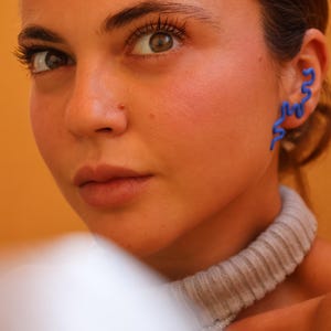 Könnte beinhalten: Ein leuchtend blauer Ear Cuff mit einem durchgehenden, wellenförmigen, schlangenförmigen Design, das der äußeren Kurve des Ohrs folgt. Das Accessoire hat eine glatte, matte Oberfläche und ist entlang des Knorpels positioniert, was einen Akzent setzt.