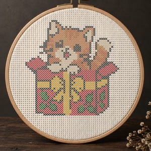 Puede incluir: Un bordado de punto de cruz en un bastidor de madera circular que muestra un gatito naranja y blanco asomándose de un regalo rojo con un lazo amarillo y detalles de hojas de acebo verdes, sobre tela blanca con un patrón de cuadrícula.