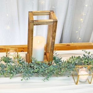 May include: Three wooden candle holders with white candles, two small gold candle holders with white candles, and a garland of green eucalyptus leaves on a white tablecloth.