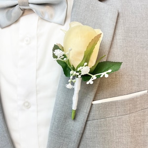 May include: A single ivory rose boutonniere with white baby's breath and green leaves. The boutonniere is pinned to a gray suit jacket. The Brides Bouquet.com is printed on the bottom right corner of the image.