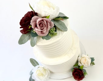 May include: Two-tiered white wedding cake decorated with white, blush pink, and burgundy artificial flowers. The cake is on a white cake stand.