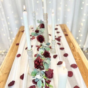May include: A white tablecloth with a garland of burgundy roses and greenery down the center.  There are five white candles in wooden candle holders along the garland.  The tablecloth is on a wooden table with burgundy rose petals scattered on the tablecloth.  The text "The Brides Bouquet.com" is in the upper right corner of the image.