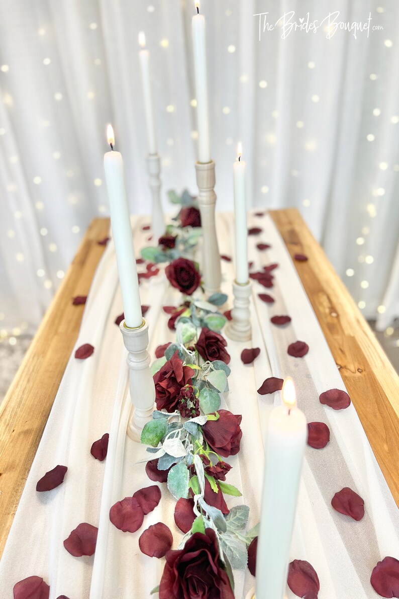 May include: A white tablecloth with a garland of burgundy roses and greenery down the center.  There are five white candles in wooden candle holders along the garland.  The tablecloth is on a wooden table with burgundy rose petals scattered on the tablecloth.  The text "The Brides Bouquet.com" is in the upper right corner of the image.