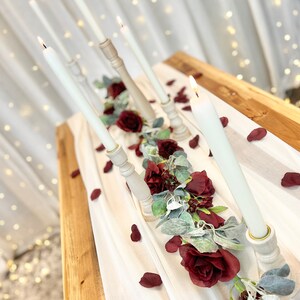 May include: A white tablecloth with a garland of burgundy roses and greenery, accented with white candles in wooden candle holders. Scattered burgundy rose petals decorate the table.