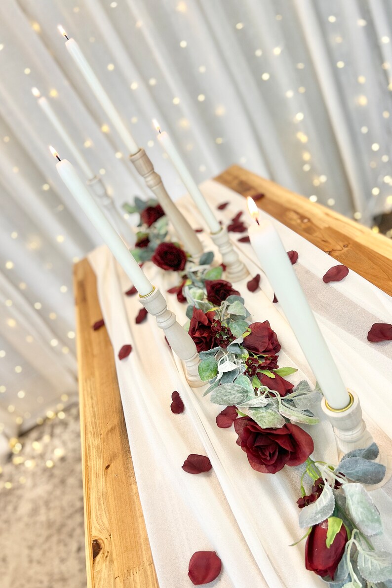 May include: A white tablecloth with a garland of burgundy roses and greenery, accented with white candles in wooden candle holders. Scattered burgundy rose petals decorate the table.