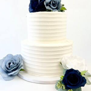 May include: Two-tiered white wedding cake decorated with blue and white roses. The cake is on a white cake stand. The cake is decorated with a simple, elegant design.