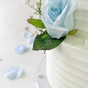 以下が含まれることがあります： 青いバラとカスミソウの花で飾られた白いウェディングケーキ。ケーキは透明なケーキスタンドの上に置かれています。