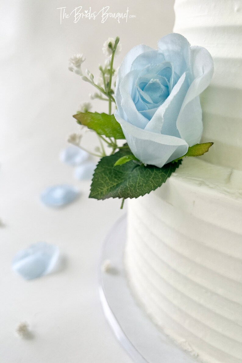 以下が含まれることがあります： 青いバラとカスミソウの花で飾られた白いウェディングケーキ。ケーキは透明なケーキスタンドの上に置かれています。