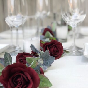 May include: A table runner with a garland of burgundy roses and green leaves. The garland is on a white tablecloth with white plates and wine glasses.