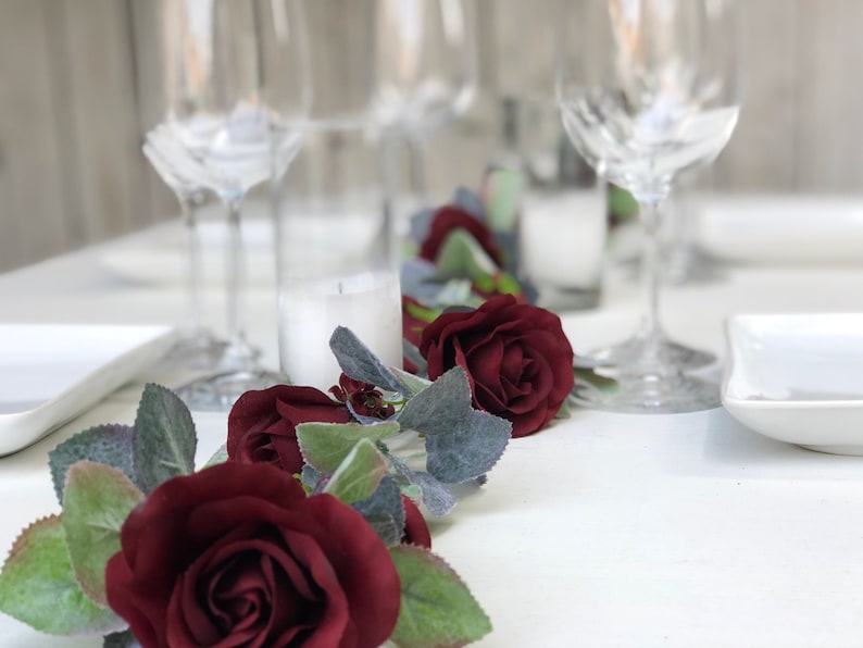 May include: A table runner with a garland of burgundy roses and green leaves. The garland is on a white tablecloth with white plates and wine glasses.