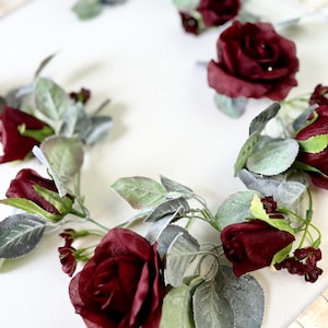 May include: A garland of burgundy roses and green leaves with a frosted finish. The garland is perfect for adding a touch of elegance to any space.