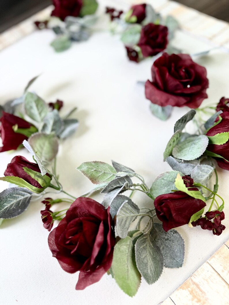 May include: A garland of burgundy roses and green leaves with a frosted finish. The garland is perfect for adding a touch of elegance to any space.