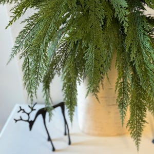 May include: A green artificial cedar branch in a white birch bark vase. The branch is hanging over the edge of the vase and has a natural, realistic look.