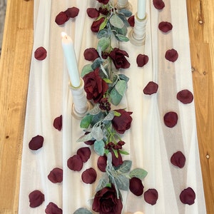 May include: A white table runner with a garland of burgundy roses and eucalyptus leaves. The runner is decorated with burgundy rose petals and three white candles in wooden candle holders.