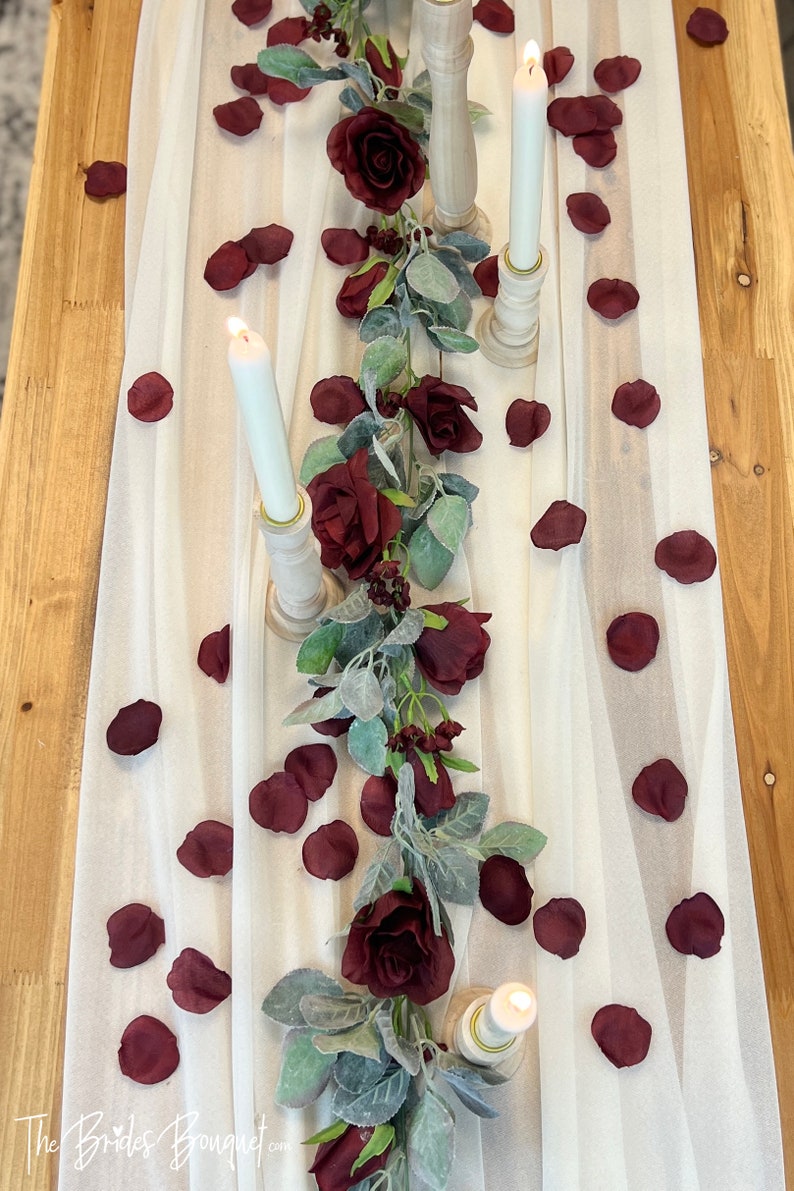 May include: A white table runner with a garland of burgundy roses and eucalyptus leaves. The runner is decorated with burgundy rose petals and three white candles in wooden candle holders.