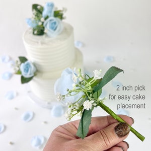 以下が含まれることがあります： 薄い青色のシルクの花束に、白いカスミソウと緑の葉が添えられています。花束は、ケーキに簡単に置くことができるように、5cmのピックに取り付けられています。