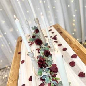 May include: White candles in wooden candle holders on a white tablecloth with burgundy rose petals and greenery.