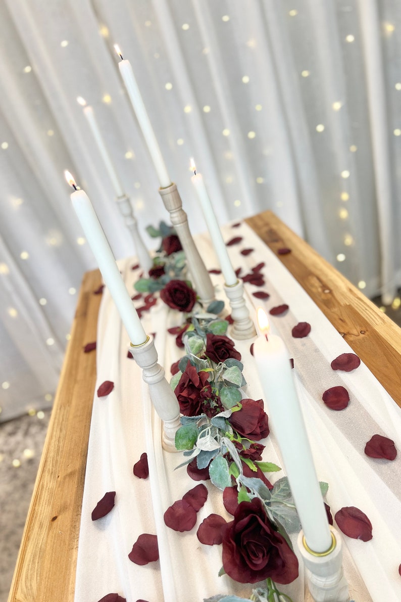 May include: White candles in wooden candle holders on a white tablecloth with burgundy rose petals and greenery.