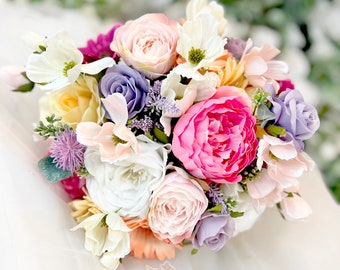 May include: A bouquet of pastel-colored artificial flowers, including pink, white, yellow, and purple blooms. The bouquet is arranged in a circular shape and features a variety of flower types, including roses, peonies, and lilies.