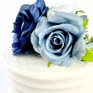 May include: A white cake with blue and navy blue fabric roses on top. The cake is decorated with white frosting and has a subtle ripple pattern.