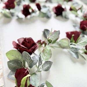 May include: A burgundy floral garland with small red roses and green leaves. The garland is arranged in a circle shape on a white background.