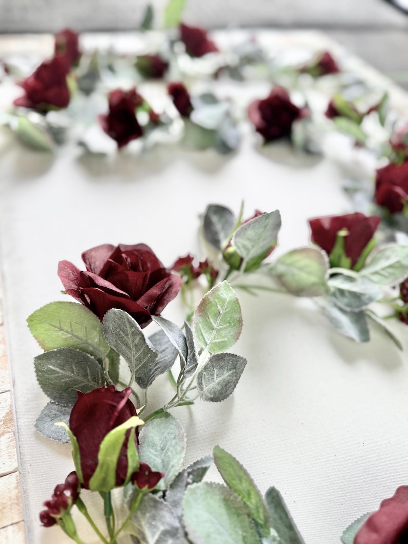 May include: A burgundy floral garland with small red roses and green leaves. The garland is arranged in a circle shape on a white background.