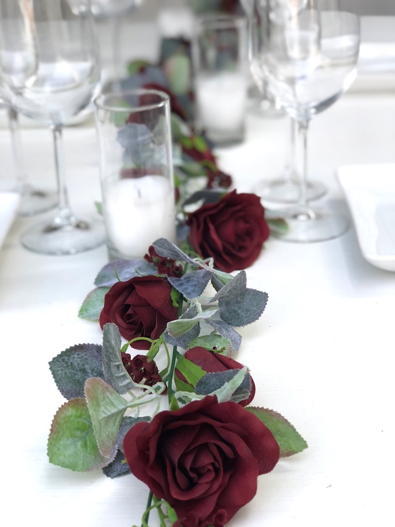 May include: A burgundy rose garland with green leaves and silver-grey foliage. The garland is on a white table with clear glass candle holders and wine glasses.