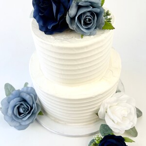 May include: Two-tiered white wedding cake decorated with blue and white roses. The cake is on a white cake stand.