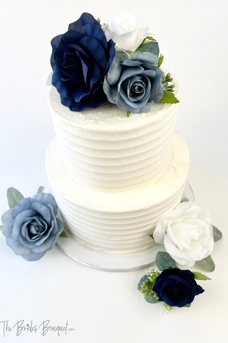May include: Two-tiered white wedding cake decorated with blue and white roses. The cake is on a white cake stand.