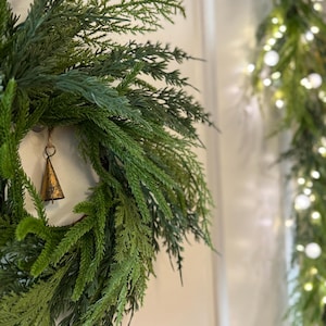 May include: A close-up of a green and blue artificial wreath with a small gold bell hanging from it. The wreath is made of faux evergreen branches. A garland with white lights is in the background.
