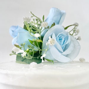 以下が含まれることがあります： 白いケーキの上に、薄い青色のバラとカスミソウの花の飾り付けが施されています。ケーキは白いバタークリームで覆われています。