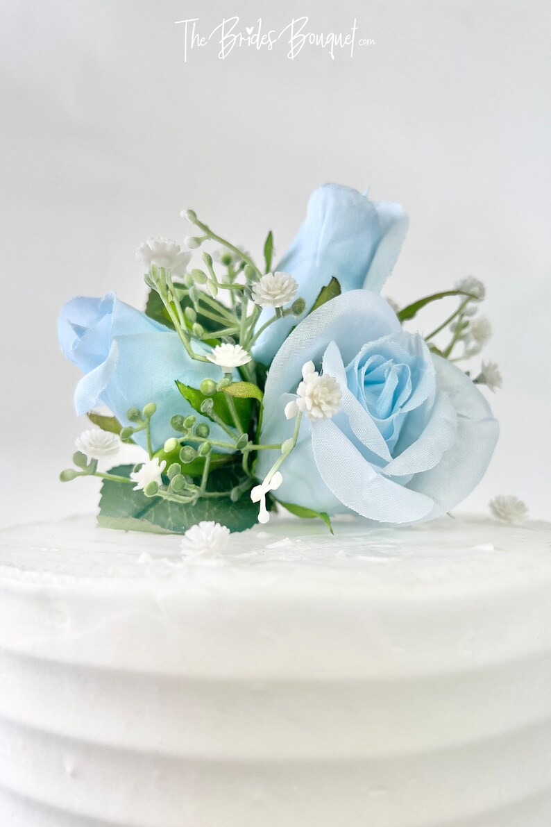 以下が含まれることがあります： 白いケーキの上に、薄い青色のバラとカスミソウの花の飾り付けが施されています。ケーキは白いバタークリームで覆われています。