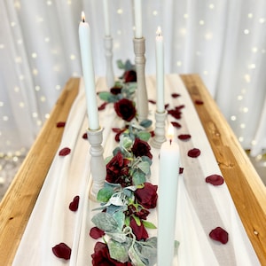 May include: A white tablecloth with a garland of burgundy roses and greenery down the center. Five white candles are in wooden candle holders along the garland. Burgundy rose petals are scattered on the tablecloth. The text "The Brides Bouquet.com" is in the top left corner of the image.