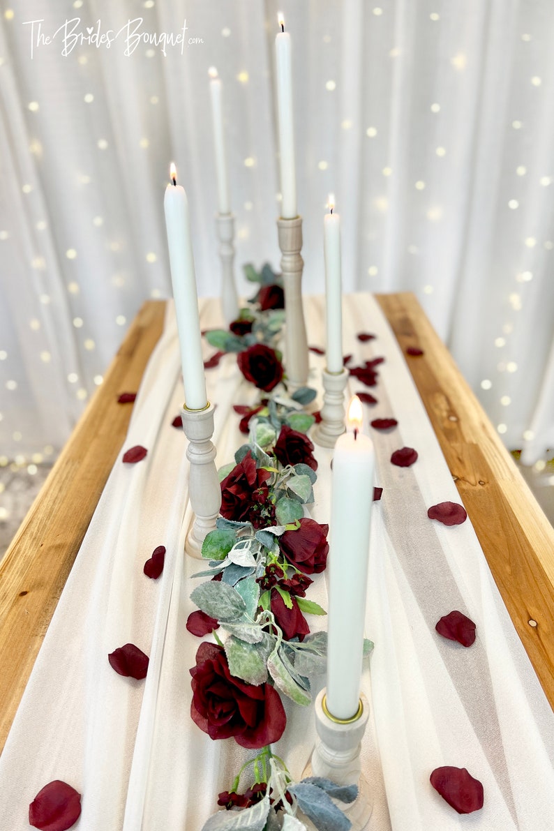 May include: A white tablecloth with a garland of burgundy roses and greenery down the center. Five white candles are in wooden candle holders along the garland. Burgundy rose petals are scattered on the tablecloth. The text "The Brides Bouquet.com" is in the top left corner of the image.