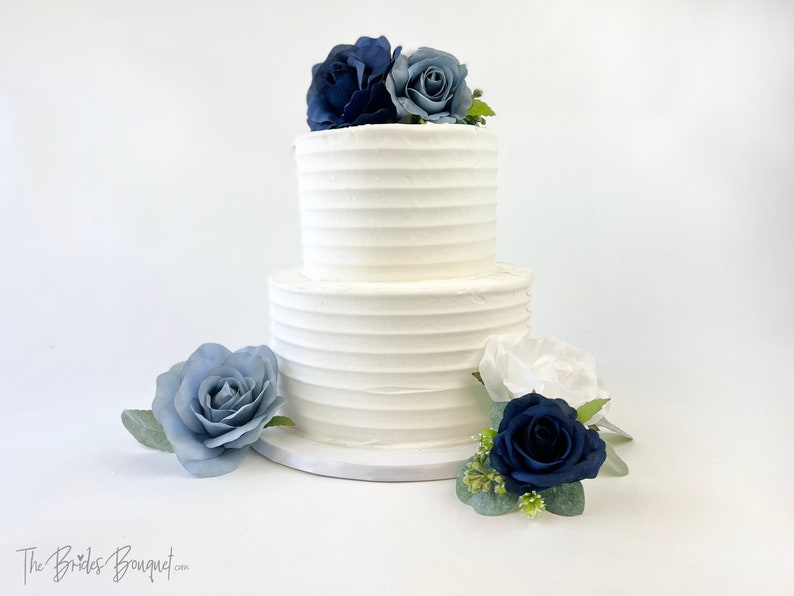 May include: Two-tiered white wedding cake decorated with blue and white roses. The cake is on a white cake stand.
