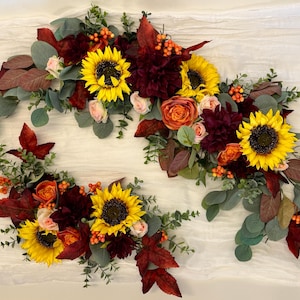 May include: Two floral arrangements featuring bright yellow sunflowers, deep burgundy dahlias, orange roses, and small orange berries. The bouquets are accented with sage green eucalyptus leaves and dark red autumn foliage, creating a seasonal display.