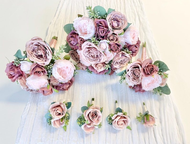 May include: A set of five bouquets of artificial flowers in shades of pink and mauve. The bouquets are arranged in a row on a white fabric background. The largest bouquet is in the center, with two smaller bouquets on either side, and two very small bouquets at the ends.