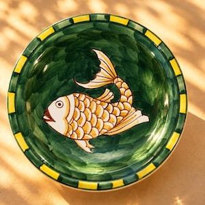 Könnte beinhalten: Eine runde Keramikschale mit einem handbemalten goldenen Fisch mit detaillierten Schuppen, der auf einem tiefgrünen Hintergrund zentriert ist. Der Rand ist mit einem sich wiederholenden Muster aus gelben und dunkelgrünen Segmenten verziert.