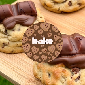 Op de afbeelding: Drie ronde koekjes met chocoladestukjes, belegd met gesmolten melkchocoladerepen. Een ronde bruine sticker met een herhalend patroon van hartvormige koekjes en de tekst THE BAKE BAR is over de koekjes geplaatst en bedekt de middelste gedeeltelijk.