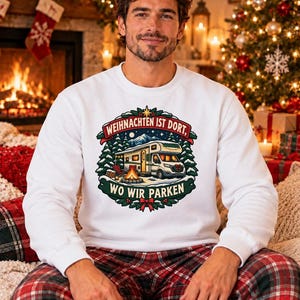 Könnte beinhalten: Ein weißes Langarm-Sweatshirt mit einem Grafikdruck eines Wohnmobils, das in einem verschneiten Wald an einem Lagerfeuer steht. Der Text WEIHNACHTEN IST DORT, WO WIR PARKEN ist von einem kreisförmigen Kranz aus Tannenzweigen und einer roten Schleife umgeben.