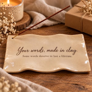 Op de afbeelding: Een rechthoekige, golvende wierookhouder van beige keramiek met de zwarte tekst: Your words, made in clay. Some words deserve to last a lifetime. Een bruin wierookstokje is in een klein gaatje aan de linkerkant van de houder gestoken.
