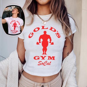 May include: A white short-sleeved t-shirt featuring a red graphic of a bodybuilder holding dumbbells. The text "GOLD'S GYM SoCal" is printed in red on the front. The shirt has a crew neckline and a fitted silhouette.