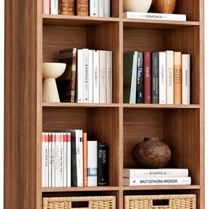 May include: A tall, eight-compartment wooden bookshelf with a walnut finish. The shelves contain various books, decorative ceramic vases, and two woven wicker storage baskets at the bottom. The books include titles like Kinfolk and Modern Architecture.