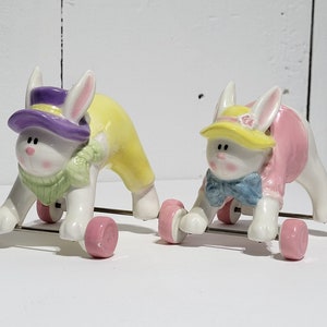 Könnte beinhalten: Zwei Keramik-Hasenfiguren auf Rädern. Der Hase links trägt einen lila Hut, einen grünen Schal und eine gelbe Hose. Der Hase rechts trägt einen gelben Hut, eine blaue Schleife und ein rosa Kleid. Beide Hasen haben rosa Räder.