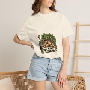 May include: A cream-colored short-sleeve t-shirt featuring a graphic of a garden fork and potatoes in soil with green leaves. The text "DIG HERE FOR DINNER" is printed in black letters on a banner below the potatoes, centered on the chest.