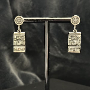 May include: A pair of silver-toned drop earrings featuring a circular stud top with a stylized face design. Each earring has a rectangular pendant hanging from the stud, embossed with a detailed figure wearing an elaborate headdress and attire.