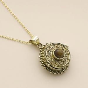 May include: A gold-toned circular locket pendant on a matching delicate chain. The locket features an intricate embossed floral pattern and a central round brown tiger's eye gemstone. The edges of the pendant are decorated with small beads.