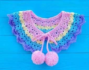 May include: A rainbow crochet collar with pink pom-poms attached to the ends of the ties. The collar is on a blue wooden background.