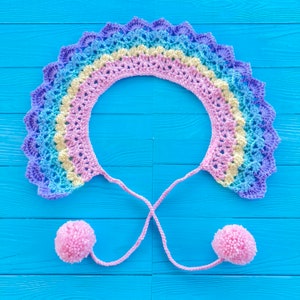 May include: A rainbow crochet collar with two pink pom-poms attached to the ends of the ties.